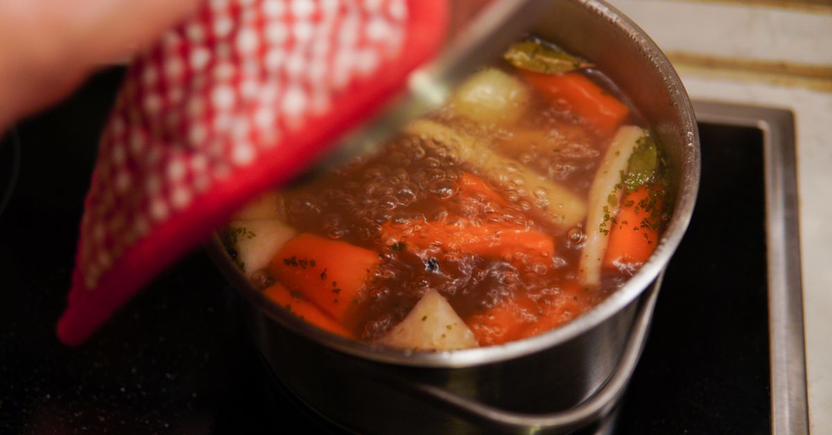 Cook lifting the lid on a pot of bones and vegetables simmering into bone broth