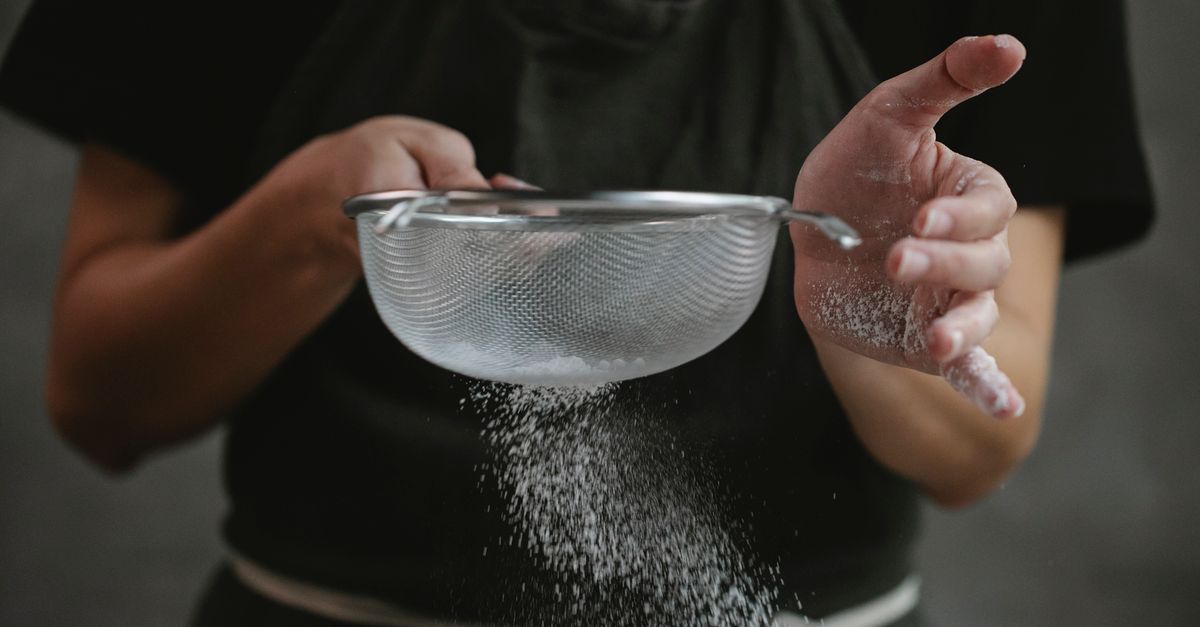 Hands sifting wheat flour through a fine sieve, the milling fineness that lifts the glycaemic index of bread