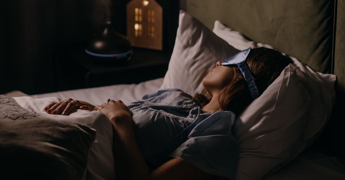 Woman asleep in a dim bedroom wearing a sleep mask, the dark sleeping environment linked to lower weight gain