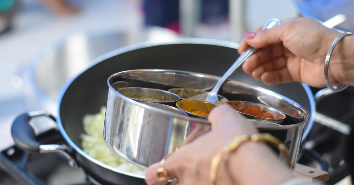 Hands cooking an Indian dish at a stove with bowls of spices beside the pan