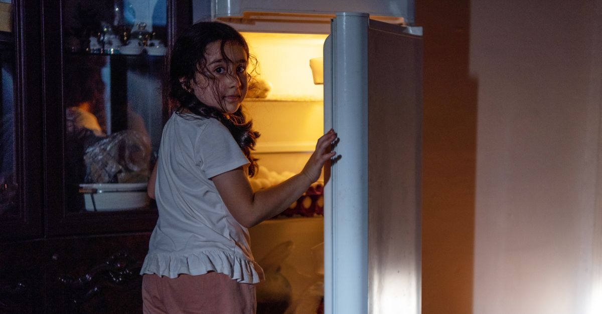Woman at an open fridge in a dark kitchen late at night, when short sleep raises evening eating