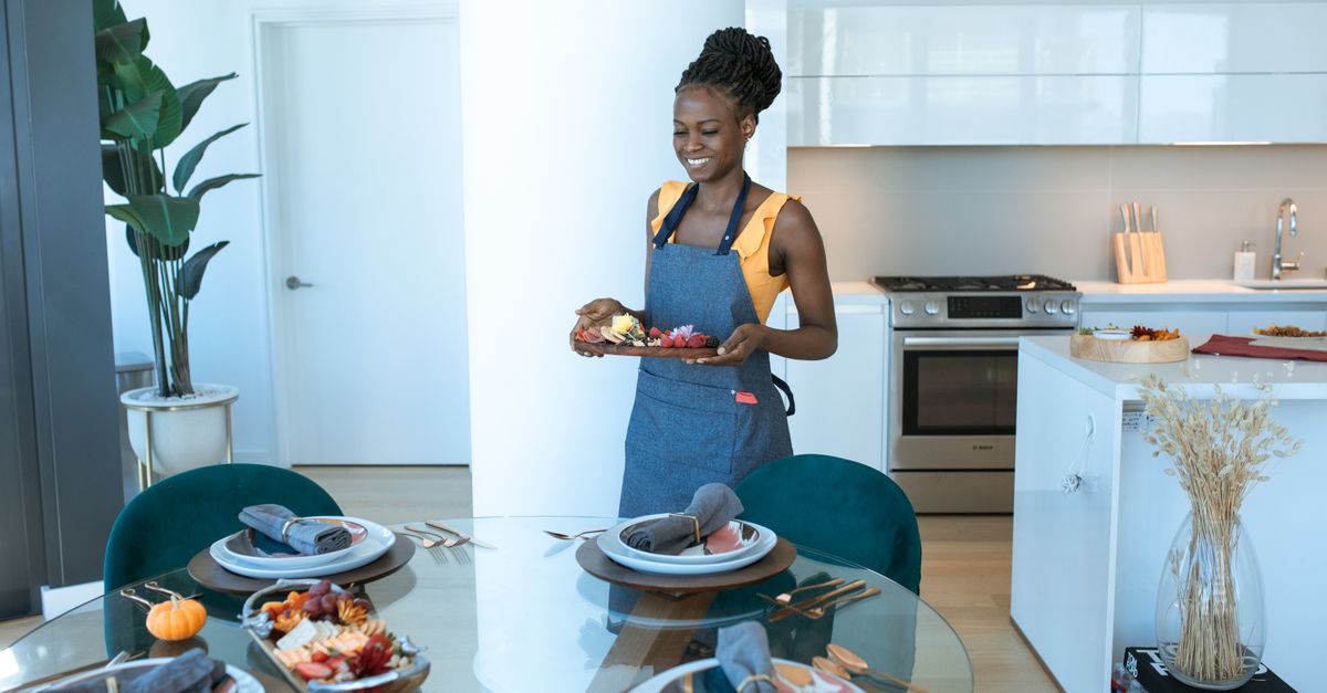 A woman in an apron setting a full plated meal on a laid kitchen table