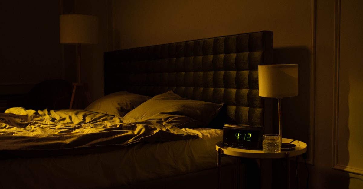 Unmade bed in a dim bedroom beside a glowing digital clock, the short sleep that worsens sugar cravings
