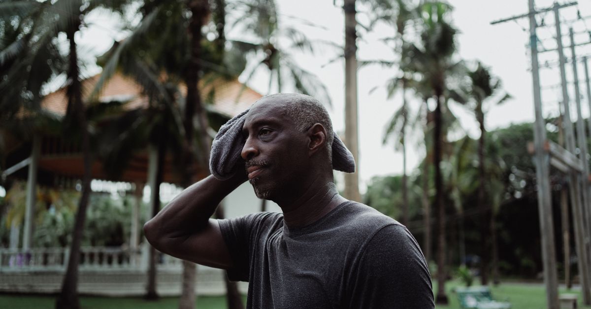 A man pauses in a park to wipe sweat from his forehead in summer heat