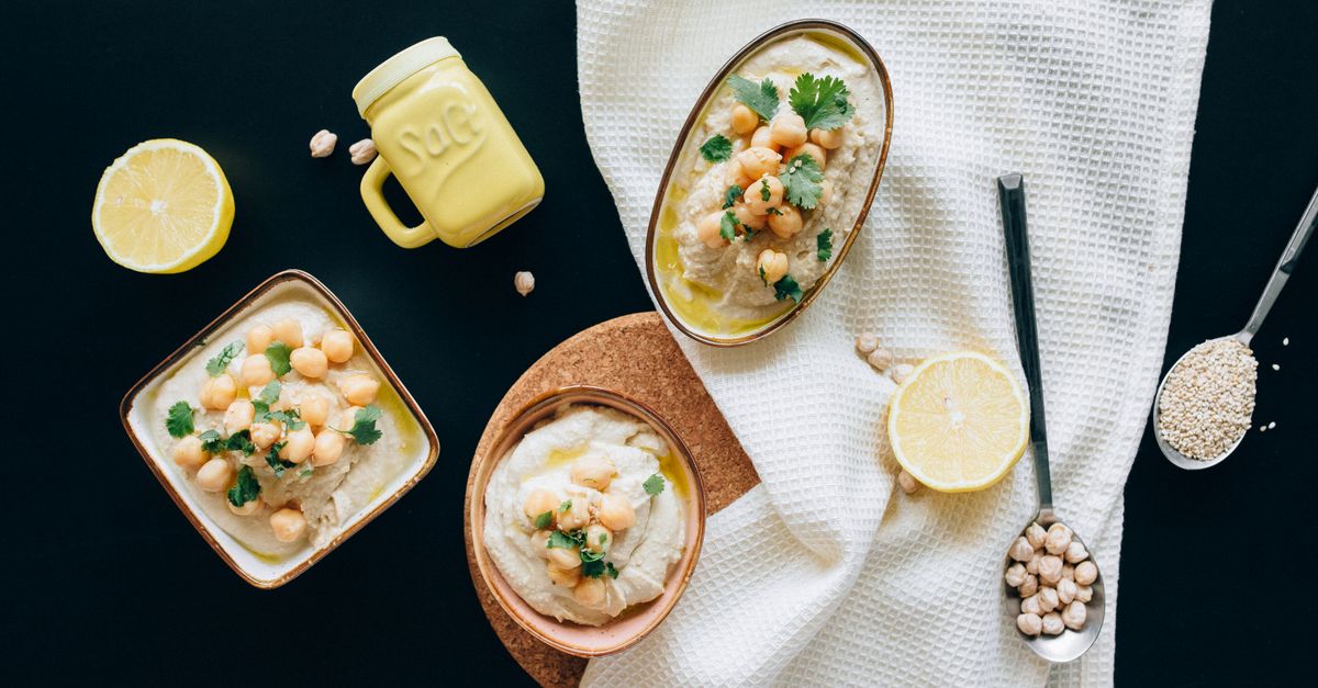 Bowls of tahini and hummus with lemon and a spoon of sesame seeds on a dark table