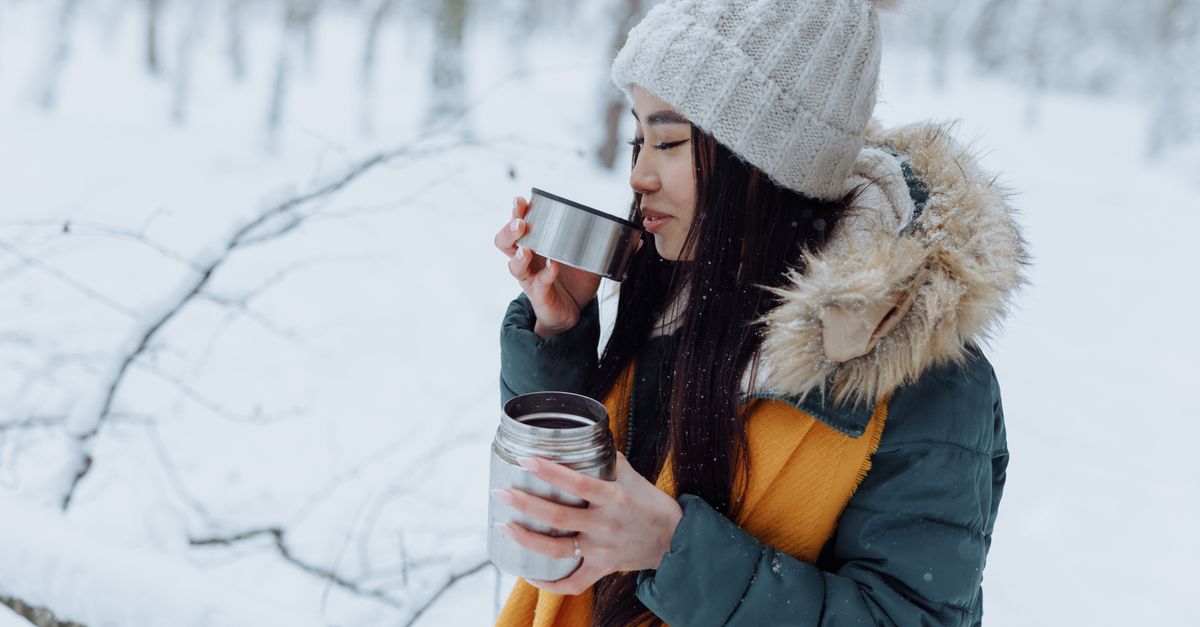 Woman in a knitted hat and scarf sipping a warm drink from a flask in a snowy forest